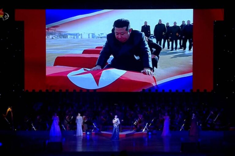 A imagem mostra um grande palco iluminado, onde várias pessoas estão se apresentando. No fundo, há uma tela grande exibindo uma cena de um homem se ajoelhando sobre uma bandeira da Coreia do Norte, com um grupo de pessoas ao fundo. Os artistas no palco estão vestidos com trajes formais e há uma orquestra visível na parte inferior da imagem. O ambiente é decorado com luzes e elementos que remetem à cultura norte-coreana.