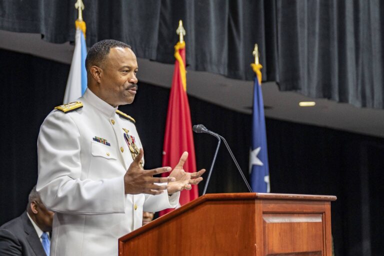 Um homem em uniforme militar branco está falando em um púlpito de madeira. Ele gesticula com as mãos enquanto se dirige a uma audiência. Ao fundo, há várias bandeiras, incluindo uma bandeira azul com estrelas. A cena parece ser de uma cerimônia formal.