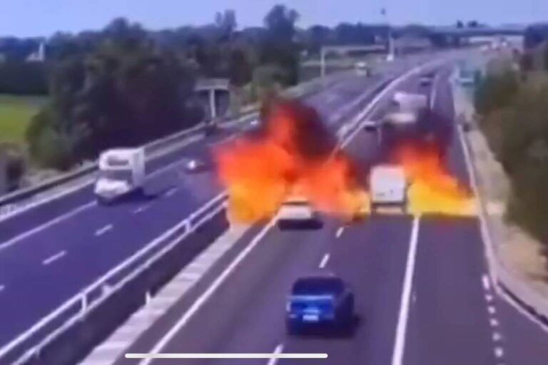 A imagem mostra uma rodovia com vários veículos. No centro da imagem, há uma grande explosão com chamas e fumaça, que se eleva em direção ao céu. À esquerda, um caminhão e, à direita, um carro azul estão visíveis. O cenário é ensolarado, com vegetação ao fundo.