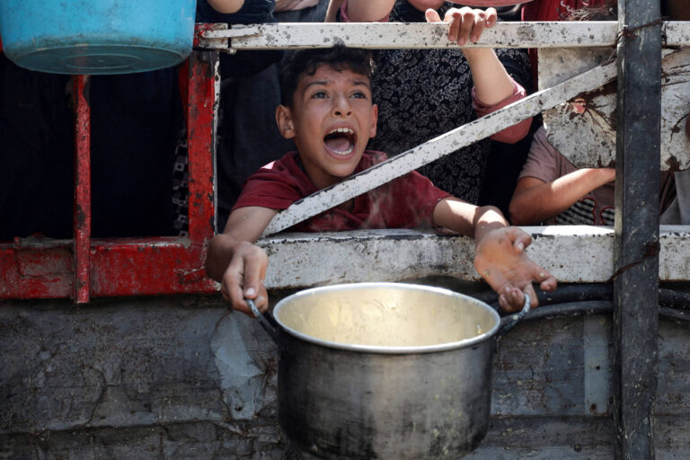 A imagem mostra uma criança com expressão de desespero, gritando enquanto segura uma panela. Ele está em um ambiente onde outras pessoas também estão visíveis, algumas delas com as mãos estendidas. O fundo é uma estrutura metálica, possivelmente uma janela ou grade, e a cena parece retratar uma situação de necessidade ou distribuição de alimentos.