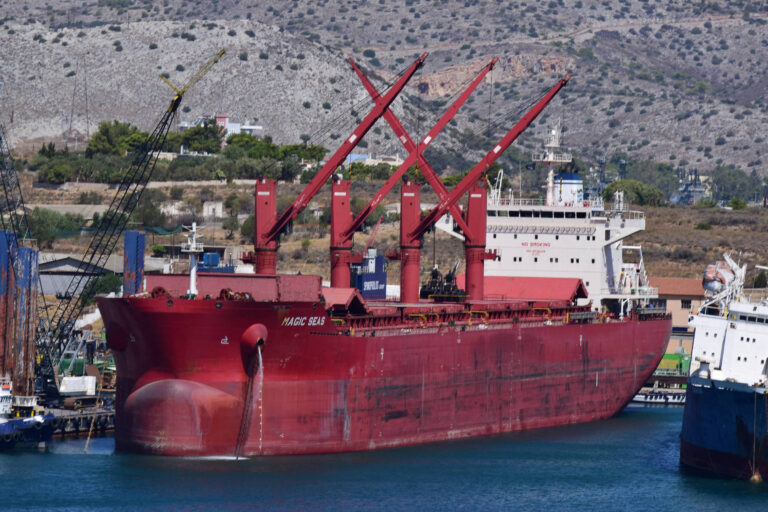 A imagem mostra um grande navio de carga vermelho ancorado em um porto. O navio possui três guindastes altos e está cercado por outros barcos e estruturas portuárias. Ao fundo, há montanhas e vegetação, indicando uma área costeira.