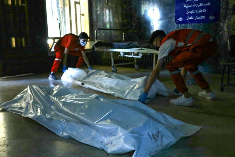 Dois socorristas estão manuseando sacos de corpo brancos em um ambiente hospitalar. O chão é de um material claro e há uma maca ao fundo. As paredes parecem desgastadas e há uma placa visível em árabe ao fundo, indicando informações sobre o local. Os socorristas estão usando uniformes vermelhos e máscaras de proteção.