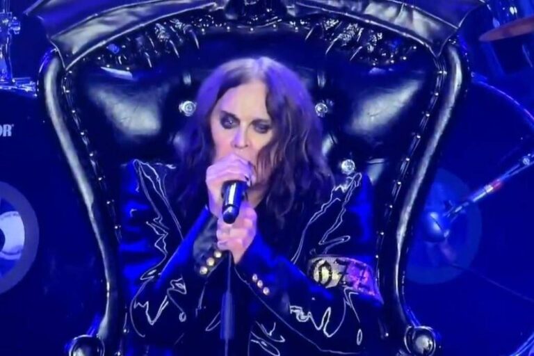 Um artista está se apresentando em um palco, sentado em uma cadeira alta e ornamentada, com um design de couro preto e detalhes em metal. Ele segura um microfone e canta, com um olhar intenso. Ao fundo, há instrumentos musicais, incluindo tambores, que fazem parte da banda. A iluminação do palco é dramática, destacando o artista e criando uma atmosfera envolvente.