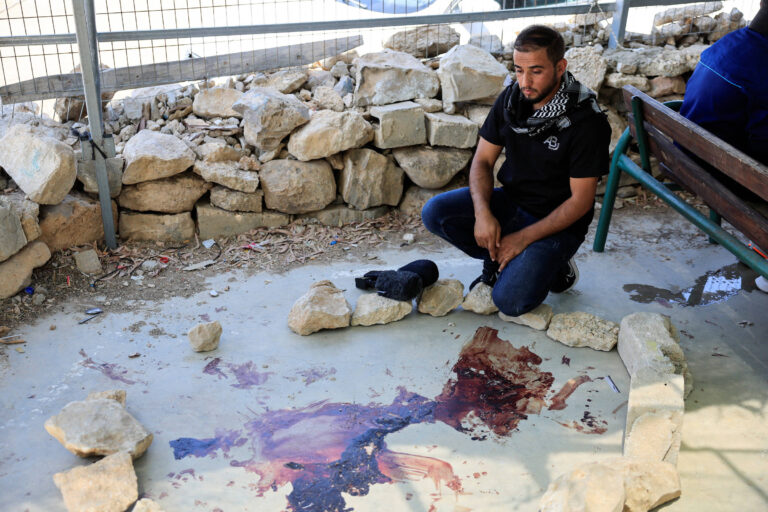 Um homem está sentado em uma área cercada por pedras, com um padrão de manchas de sangue no chão. Ele está usando uma camiseta escura e um lenço ao redor do pescoço. O ambiente parece ser ao ar livre, com uma grade ao fundo e pedras empilhadas ao redor. O homem parece pensativo e em luto.