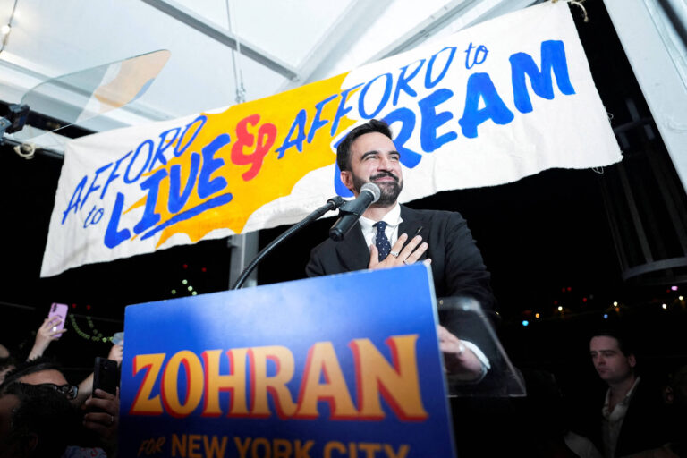 Um homem em um palanque fala para uma multidão em um evento político. Ele está vestido com um terno escuro e sorrindo, com a mão sobre o coração. Atrás dele, há um banner grande que diz