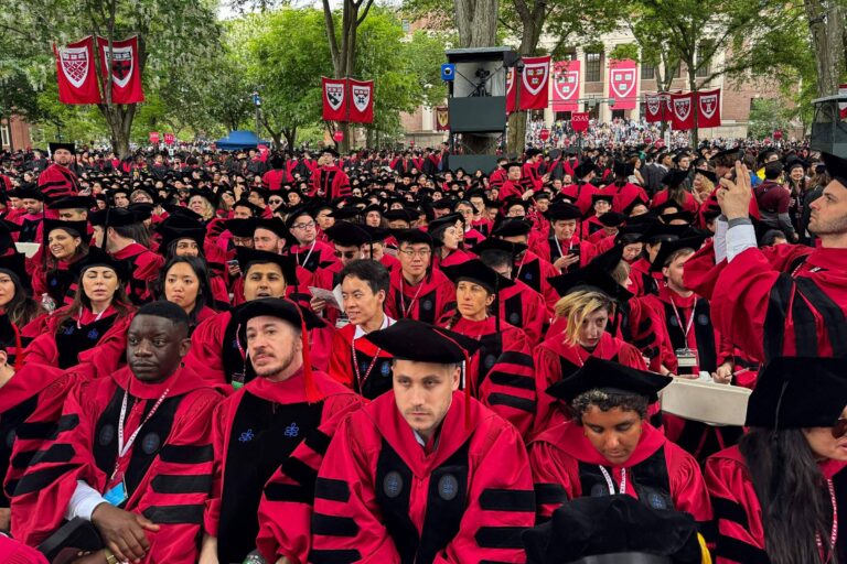 A imagem mostra uma grande multidão de graduados sentados em um evento de formatura. Eles estão vestidos com togas e chapéus de formatura, predominantemente em vermelho e preto. Ao fundo, há bandeiras de uma instituição de ensino, possivelmente Harvard, e árvores ao redor. A atmosfera é de celebração e solenidade, com muitos graduados olhando para frente e alguns interagindo entre si.