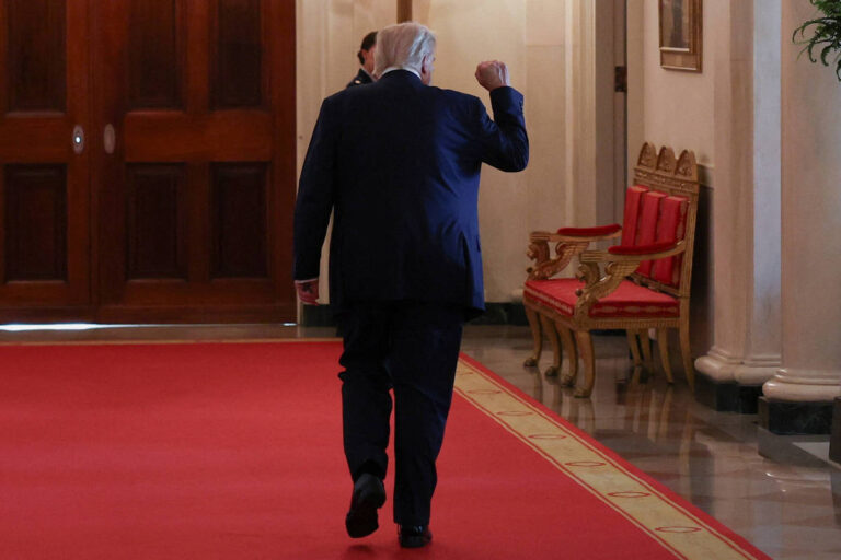 Um homem de terno escuro está caminhando em um corredor com um tapete vermelho. Ele está de costas, levantando o punho em um gesto de celebração ou determinação. Ao fundo, há portas de madeira e algumas cadeiras vermelhas dispostas ao longo da parede.