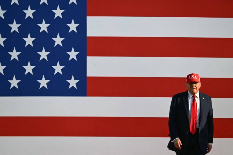 Um homem está em pé em frente a uma grande bandeira dos Estados Unidos. Ele usa um chapéu vermelho com a inscrição