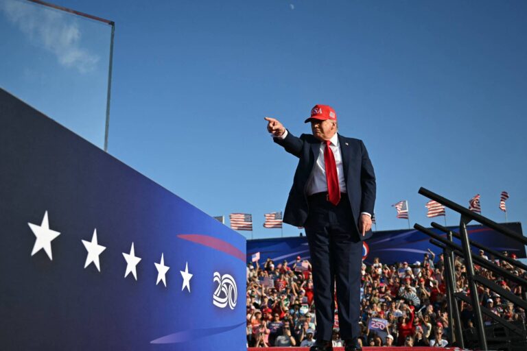 Um homem vestido com um terno escuro e uma gravata vermelha, usando um boné vermelho, está em um palco apontando para a frente. Ao fundo, há uma multidão de pessoas e bandeiras dos Estados Unidos. O cenário é ensolarado e claro, com um céu azul.