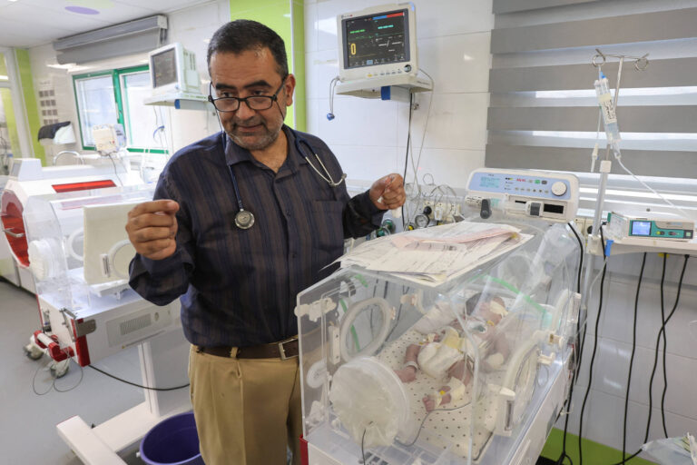 Um médico, usando óculos e um estetoscópio, está em pé ao lado de uma incubadora com um recém-nascido dentro. O ambiente é uma unidade neonatal, com equipamentos médicos visíveis ao fundo, incluindo monitores e dispositivos de suporte à vida. O médico parece estar revisando documentos enquanto observa o bebê na incubadora.