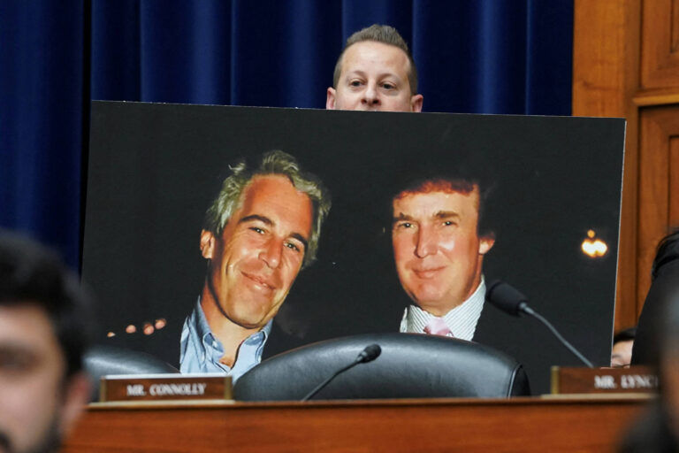 A imagem mostra um homem segurando um grande retrato de duas pessoas em uma audiência. O fundo é uma sala com uma cortina azul e mesas de audiência. O homem que segura o retrato está parcialmente visível, enquanto o retrato exibe duas figuras masculinas sorrindo. A mesa à frente tem placas com nomes, e há outras pessoas visíveis na imagem, mas não estão claramente focadas.