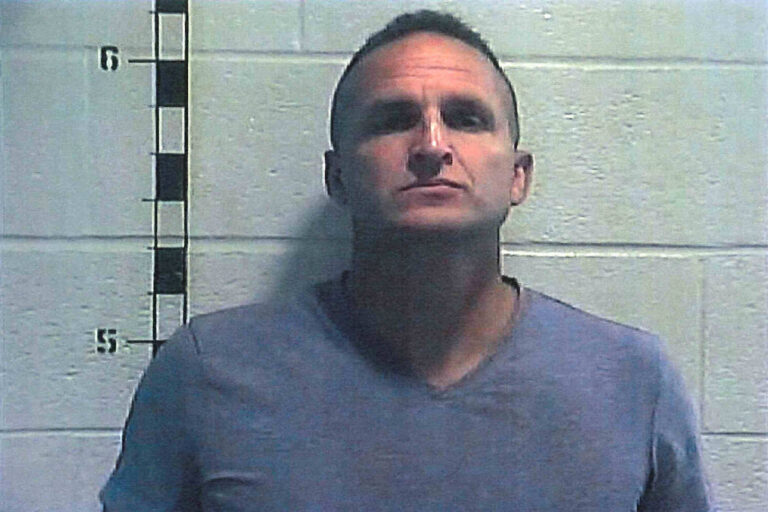 A imagem mostra um homem em um fundo de parede de tijolos claros. Ele está de pé, olhando diretamente para a câmera, com uma expressão séria. O homem usa uma camiseta cinza e tem cabelo curto. À esquerda da imagem, há uma régua de altura marcada em centímetros, indicando a altura do indivíduo. A iluminação é suave, e a imagem parece ser uma foto de registro policial.