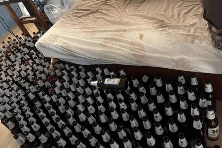 A imagem mostra um quarto com uma cama desfeita e coberta por um lençol. Ao redor da cama, há uma grande quantidade de garrafas dispostas em fileiras, com os rótulos visíveis. No fundo, pode-se ver uma mesa com um computador e outros objetos. A iluminação é natural, proveniente de janelas com cortinas.