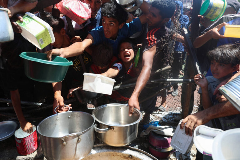 A imagem mostra um grupo de pessoas, principalmente crianças e jovens, em uma situação de grande agitação enquanto tentam pegar alimentos. Eles estão se estendendo para frente, segurando recipientes como panelas e tigelas, em um ambiente ao ar livre. O cenário parece ser de uma distribuição de alimentos, com uma grande panela ao centro, e as pessoas estão muito próximas umas das outras, demonstrando a urgência da situação.