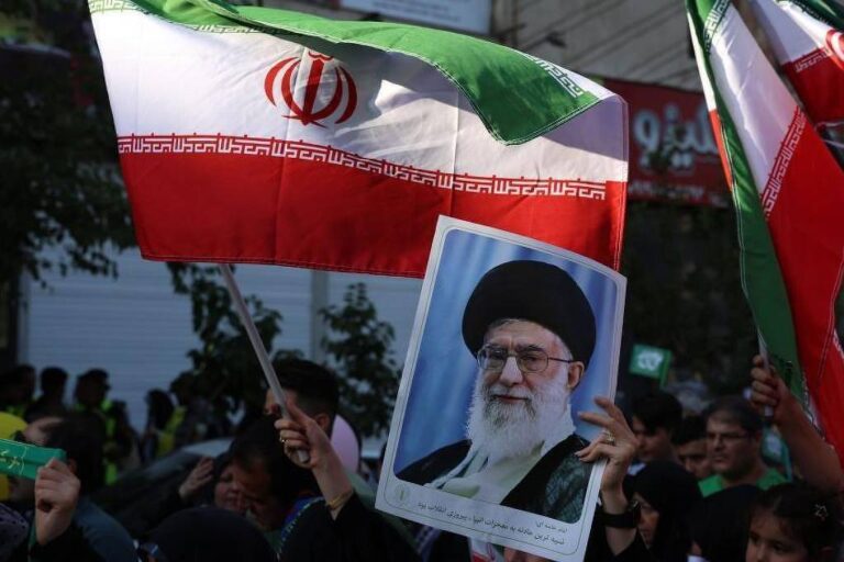 A imagem mostra um grupo de pessoas em um protesto, segurando bandeiras do Irã. Algumas pessoas também estão segurando um retrato de um líder iraniano. O ambiente parece ser urbano, com edifícios ao fundo e uma atmosfera de manifestação.