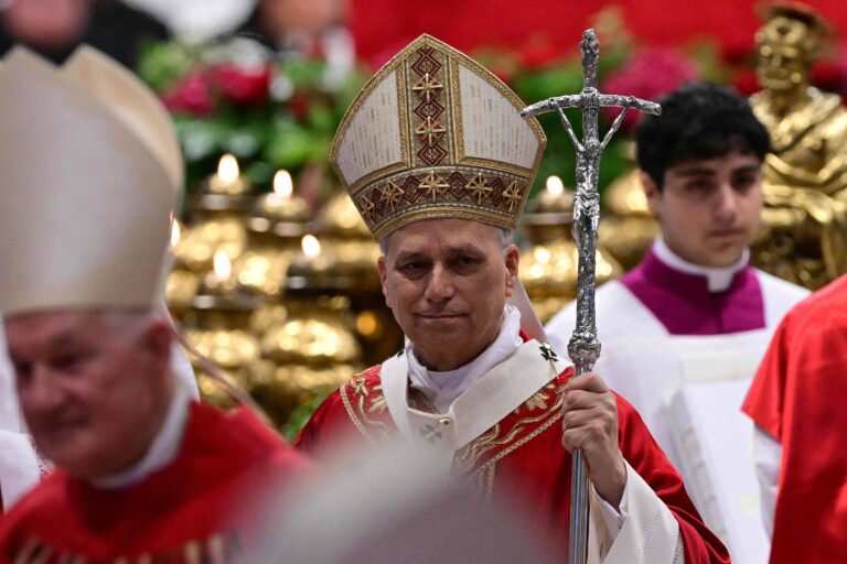 A imagem mostra um grupo de clérigos em uma cerimônia religiosa. Um dos clérigos, que está em destaque, usa um manto vermelho e um chapéu alto, segurando um bastão com uma cruz. Ao fundo, há outros clérigos e uma decoração elaborada com flores e velas. A atmosfera é solene e festiva, típica de celebrações religiosas.