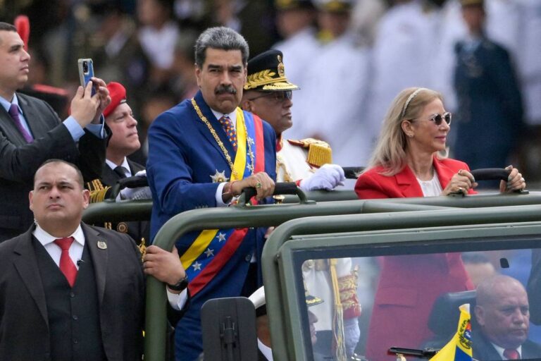 A imagem mostra um grupo de pessoas em um evento oficial ao ar livre. No centro, um homem com um uniforme militar e uma faixa colorida, que parece ser uma figura de autoridade, está em um veículo aberto. Ao seu lado, uma mulher com cabelo longo e liso, vestindo um blazer vermelho, observa o evento. Ao fundo, há várias pessoas em trajes formais, incluindo militares e civis, que assistem à cerimônia.