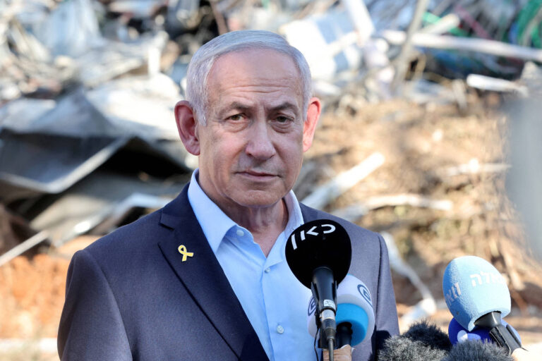 Um homem de cabelo grisalho e terno escuro está em pé em frente a um fundo de destroços. Ele parece estar falando para a imprensa, com microfones de diferentes emissoras de televisão à sua frente. O homem usa uma camisa clara e um broche amarelo na lapela. O ambiente ao fundo é de uma área devastada, com materiais de construção e entulho visíveis.