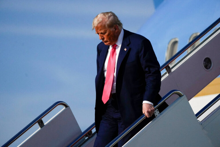 Um homem desce as escadas de um avião. Ele está usando um terno escuro e uma gravata rosa. O céu está claro e há uma parte do avião visível ao fundo.