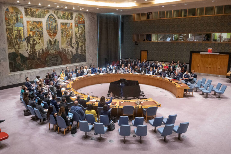 A imagem mostra uma reunião do Conselho de Segurança da ONU, com várias pessoas sentadas em torno de uma mesa em formato circular. Ao fundo, há um grande mural colorido que retrata cenas de paz e cooperação. A sala é bem iluminada e possui cadeiras azuis dispostas ao redor da mesa. Há um grande número de participantes, incluindo diplomatas e representantes de diferentes países.