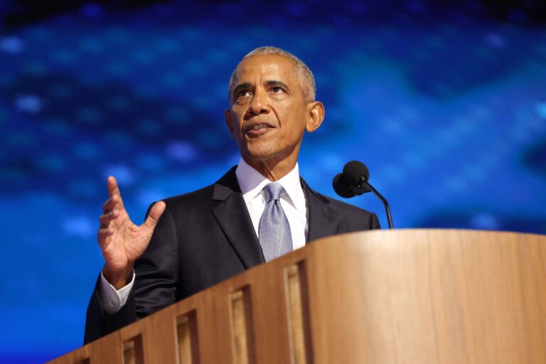Barack Obama, um homem em um terno escuro, está em pé atrás de um púlpito, gesticulando com a mão direita enquanto fala. O fundo é azul com um padrão de luzes. O homem parece estar em um evento formal, possivelmente fazendo um discurso.
