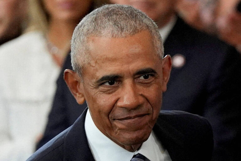 A imagem mostra um homem com cabelo curto e grisalho, usando um terno escuro e uma gravata. Ele está sorrindo levemente e parece estar em um ambiente formal, com outras pessoas ao fundo, embora não sejam claramente visíveis.