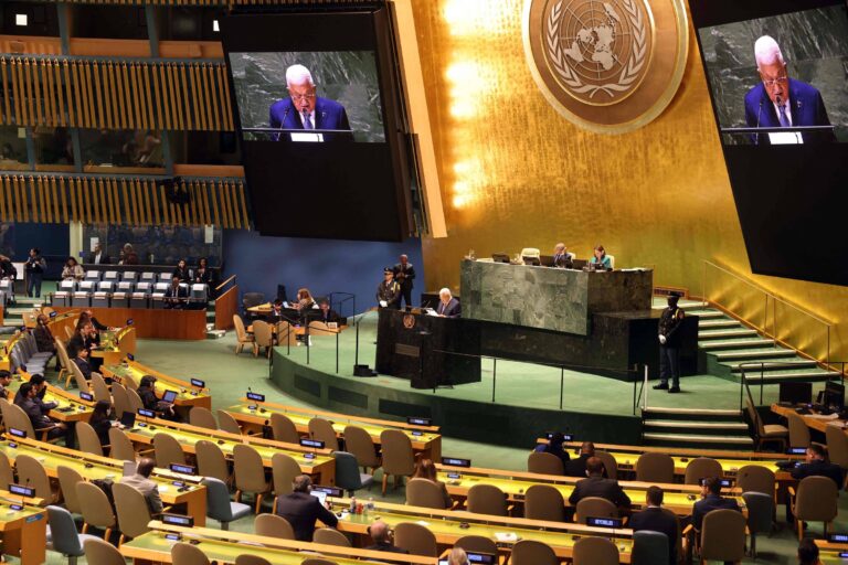 A imagem mostra uma sessão da Assembleia Geral da Organização das Nações Unidas (ONU). No centro, um orador está falando em um púlpito, enquanto uma tela grande exibe sua imagem. O auditório é amplo, com várias cadeiras ocupadas por delegados. Ao fundo, há uma grande parede com o emblema da ONU. A iluminação é suave e o ambiente é formal, refletindo a importância do evento.