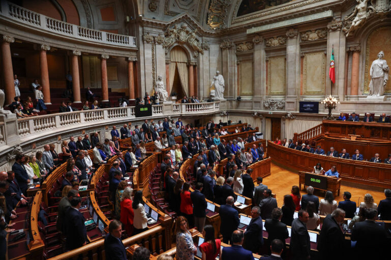 A imagem mostra o interior de um parlamento, com várias pessoas em pé e sentadas. O ambiente é grandioso, com colunas, estátuas e uma bandeira visível ao fundo. Há um painel digital que exibe um cronômetro. Os membros do parlamento estão vestidos formalmente, e a disposição das cadeiras é em formato semicircular.