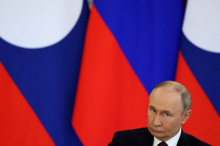 Um homem com cabelo curto e liso, usando um terno escuro e gravata, está sentado em frente a bandeiras da Rússia. As bandeiras são vermelhas, brancas e azuis, dispostas em um fundo. O homem parece estar em uma conferência ou evento formal, com uma expressão séria.