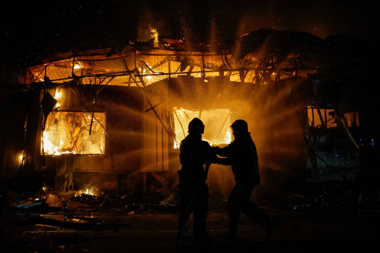 A imagem mostra um edifício em chamas, com grandes chamas e fumaça saindo das janelas. Duas pessoas, aparentemente bombeiros, estão em primeiro plano, ajudando uma à outra em meio ao incêndio. O ambiente é escuro, iluminado pelas chamas, criando um contraste dramático.
