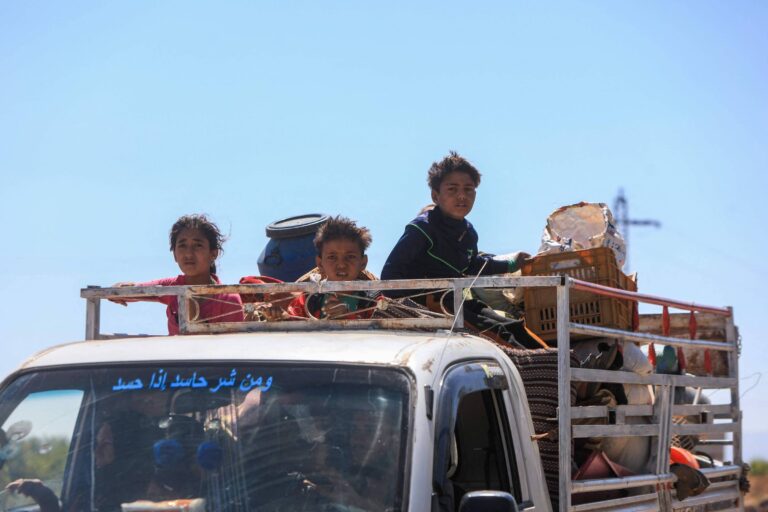 A imagem mostra um caminhão com crianças sentadas na parte de trás. Três crianças estão visíveis, duas delas são meninas e uma é um menino. Elas estão cercadas por objetos e pertences, com um céu azul ao fundo. Na parte da frente do caminhão, há uma inscrição em árabe.