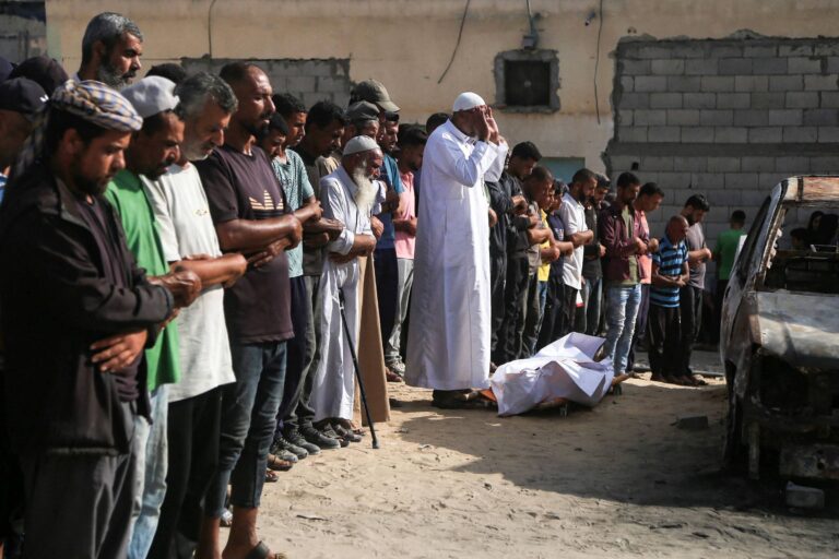 Um grupo de homens está em pé, alinhados, em uma cerimônia de luto. Eles estão com as mãos unidas em oração, alguns usando roupas tradicionais. Um homem em destaque, vestido de branco, está em posição de oração. No chão, há um corpo coberto por um lençol branco, próximo a um carro danificado. O ambiente parece ser ao ar livre, com uma parede de tijolos ao fundo.