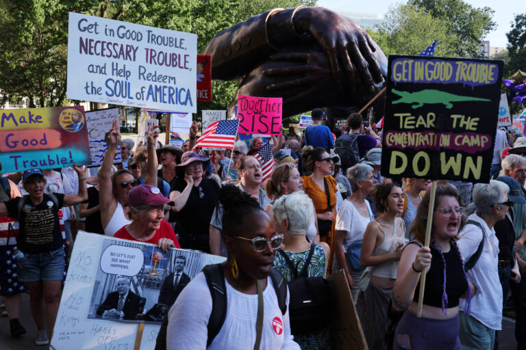 A imagem mostra um grande grupo de pessoas participando de um protesto ao ar livre. Muitas delas seguram placas com mensagens como