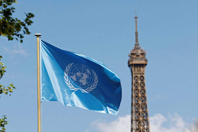 A imagem mostra uma bandeira azul da Organização das Nações Unidas (ONU) tremulando ao vento, com o símbolo da ONU em branco. Ao fundo, é visível a Torre Eiffel, um famoso monumento de Paris, sob um céu claro e ensolarado.