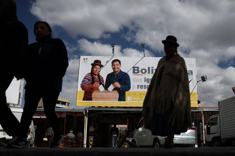 A imagem mostra um painel publicitário em uma área urbana, com duas pessoas sorrindo e segurando uma cédula de votação. O fundo é de um céu nublado e há várias pessoas caminhando em primeiro plano, algumas em silhueta. O painel contém o texto