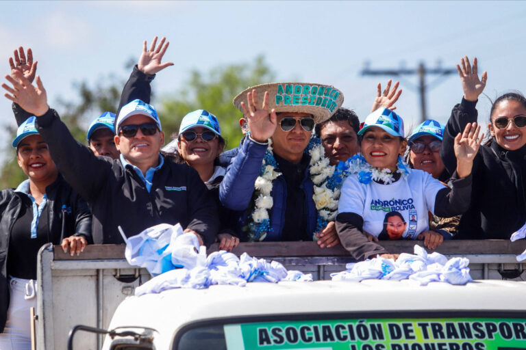 A imagem mostra um grupo de pessoas em um caminhão, acenando e sorrindo. Algumas pessoas usam camisetas e bonés azuis, enquanto outras estão vestidas com roupas escuras. Um homem no centro usa um chapéu grande e está cercado por flores. Ao fundo, há uma paisagem com árvores e postes de eletricidade. Na frente do caminhão, há uma faixa com o texto