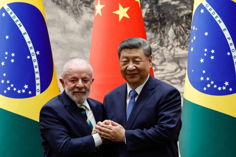 Na imagem, dois homens estão se cumprimentando em um evento oficial. À esquerda, um homem de cabelo grisalho e barba, vestindo um terno escuro e uma gravata listrada, representa o Brasil. À direita, um homem com cabelo escuro e liso, usando um terno escuro e uma gravata clara, representa a China. Ao fundo, há bandeiras do Brasil e da China, com a bandeira chinesa em destaque. O ambiente parece ser um salão de conferências.