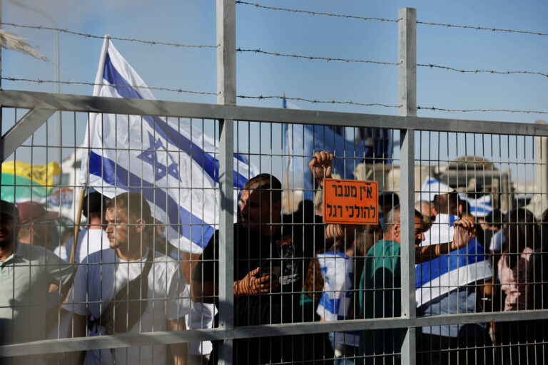 A imagem mostra um grupo de pessoas em um protesto, segurando bandeiras de Israel. Elas estão atrás de uma cerca de arame farpado. Um dos manifestantes segura um cartaz laranja com texto em hebraico. O ambiente parece ser ao ar livre, com um céu claro ao fundo.