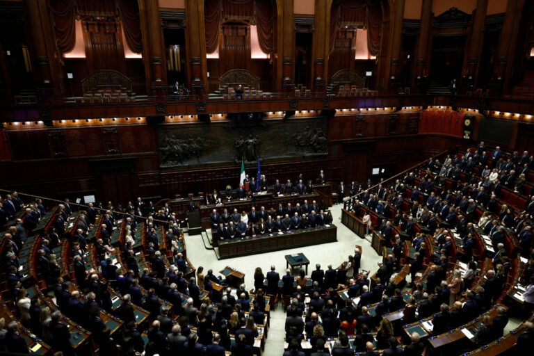 A imagem mostra uma sessão legislativa em um grande salão. Há várias fileiras de assentos ocupados por pessoas, algumas delas de pé. No centro, um grupo de pessoas está em uma mesa, possivelmente apresentando ou discutindo algo. O ambiente é formal, com decoração elaborada e bandeiras visíveis ao fundo.