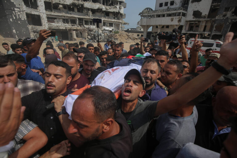 A imagem mostra um grupo de pessoas carregando um corpo coberto por um pano, em meio a uma multidão. O cenário é de destruição, com edifícios danificados ao fundo. As expressões das pessoas variam entre tristeza e agitação, e algumas estão levantando os braços. O ambiente parece ser de um funeral em uma área afetada por conflitos.