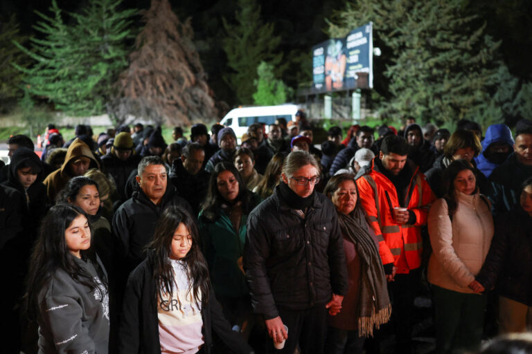 Uma multidão de pessoas está reunida em um local ao ar livre durante a noite. Algumas pessoas estão de mãos dadas, enquanto outras observam em silêncio. A maioria está vestida com roupas quentes, como casacos e gorros. Ao fundo, há uma van branca e um outdoor visível com anúncios. Árvores cercam a área, criando um ambiente natural.