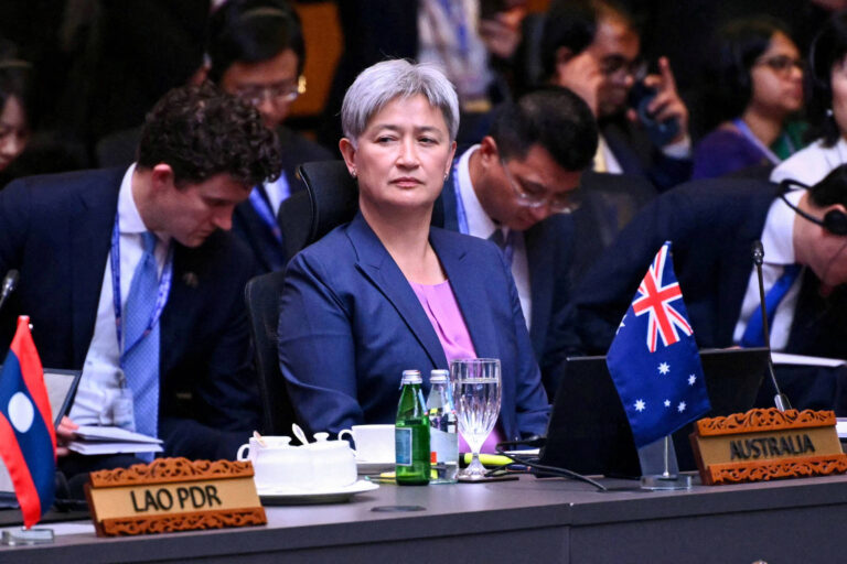 A imagem mostra uma mulher com cabelo curto e grisalho, vestindo uma blusa roxa e um blazer escuro, sentada em uma mesa de conferência. À sua frente, há uma bandeira da Austrália e uma garrafa de água. Ao fundo, outras pessoas estão sentadas, algumas olhando para baixo e outras em conversação. Também é visível uma bandeira do Laos na mesa ao lado.