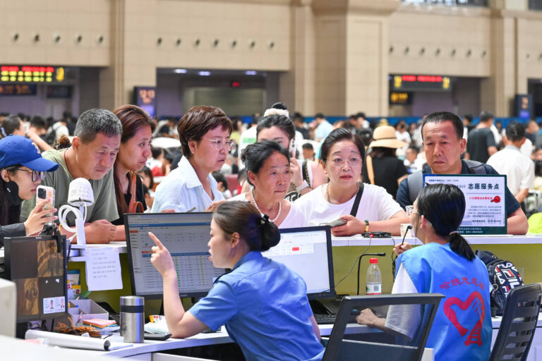 A imagem mostra um grupo de pessoas em uma estação de trem, reunidas em frente a um balcão de atendimento. Algumas pessoas estão conversando com uma funcionária que está sentada atrás do balcão, enquanto outras observam atentamente. O ambiente é movimentado, com várias pessoas ao fundo, e há painéis de informações visíveis na parte superior da imagem.