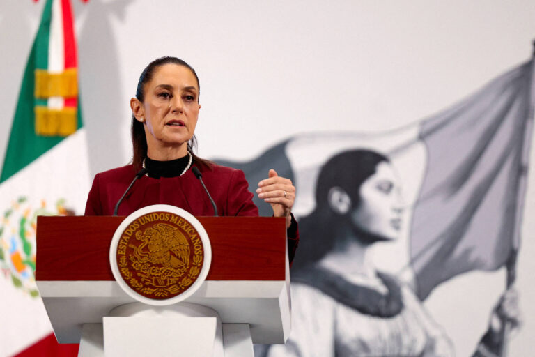 Uma mulher em pé atrás de um púlpito com o brasão do México, fazendo um discurso. Ela usa um vestido escuro e tem cabelo preso. Ao fundo, há uma imagem em preto e branco de uma mulher segurando uma bandeira. À esquerda, a bandeira do México está visível.