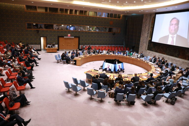 A imagem mostra uma reunião do Conselho de Segurança da ONU, com várias pessoas sentadas em cadeiras dispostas em um formato circular. Há uma tela grande exibindo a imagem de um orador. O ambiente é formal, com decoração em tons neutros e cadeiras em azul e vermelho. A sala está cheia, com muitos participantes visíveis.